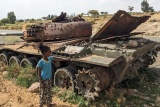 A child stands next to an abandoned tank next to a road.