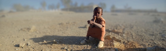 Dítě sedící na zemi na pozadí suché a vyprahlé pouštní krajiny.